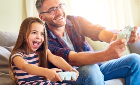 Father and daughter sitting side by side on a couch playing video games