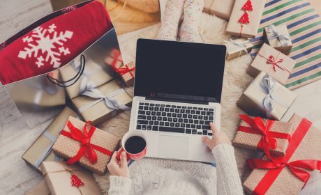 Person holding a cup of coffee whilst using their laptop surrounded by holiday gifts
