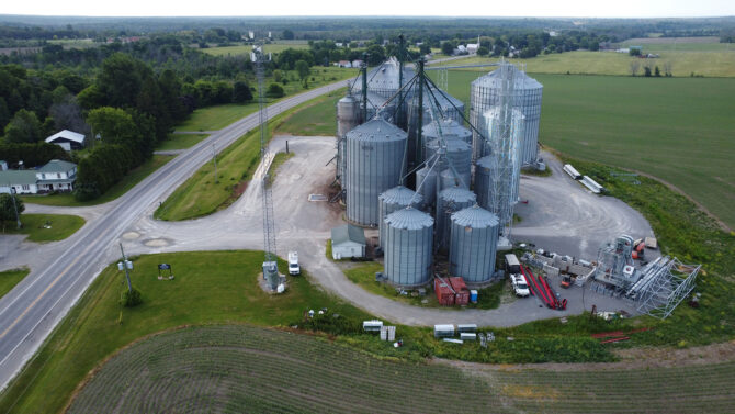 A photo taken from a drone above a large farm in a rural Ontario community surrounded by green grass and trees with many silos and grain elevators. There is an Xplore tower on the property and visible in the image.