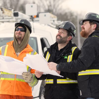 Three network technicians in helmets looking up toward fibre optic cables being installed.