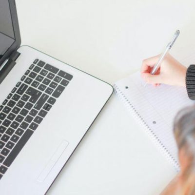 Person writing on paper whilst looking at a laptop