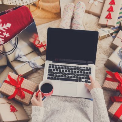 Person holding a cup of coffee whilst using their laptop surrounded by holiday gifts