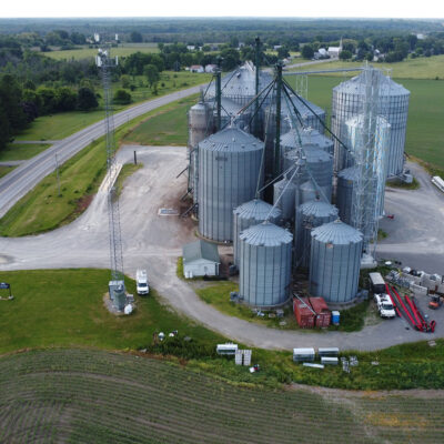 A photo taken from a drone above a large farm in a rural Ontario community surrounded by green grass and trees with many silos and grain elevators. There is an Xplore tower on the property and visible in the image.